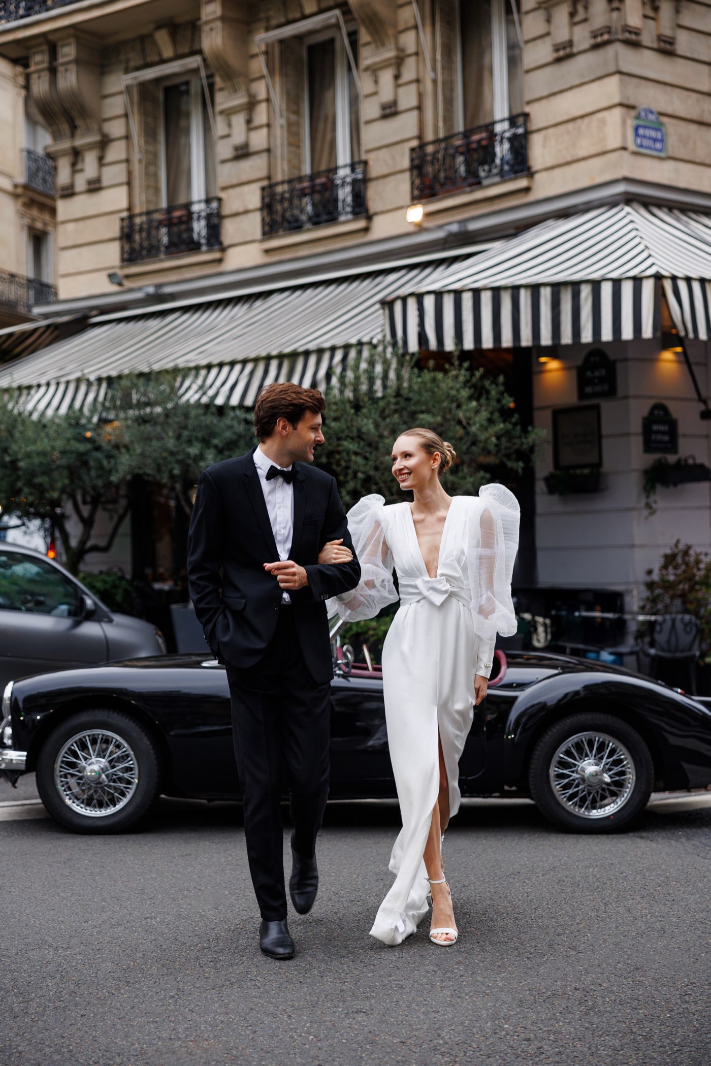 Couple walking arm in arm past a Paris café with a vintage convertible